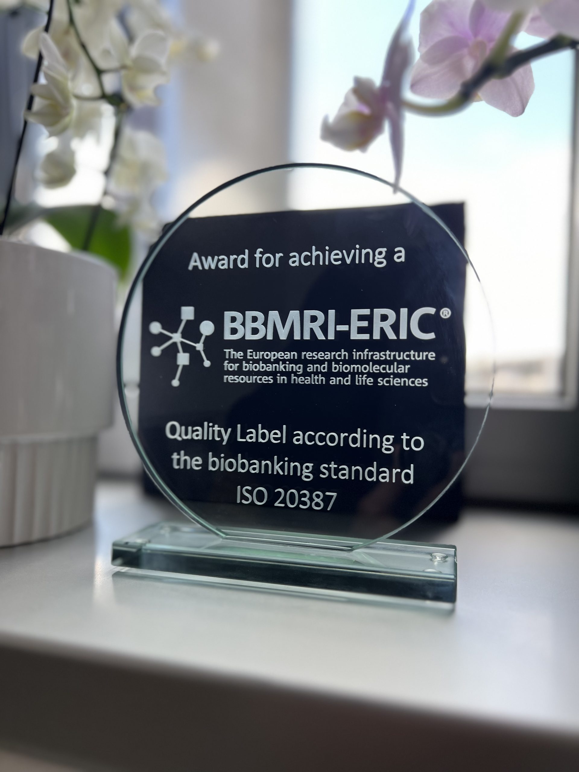 A trophy made of glass is positioned in front of a window surrounded by flowers in pots. The text on the trophy reads "BBMRI-ERIC! and " Quality Label according to the biobanking standard ISO 20387".