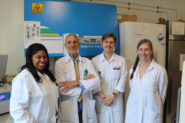 The picture of a group of four scientists, three women and a men. They are all wearing a white medical gown and smiling at the camera.