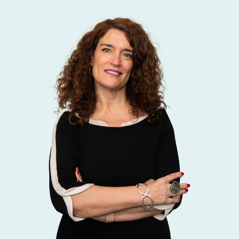 photograph of a hispanic woman with long curly dark hair who is smiling at the camera. She wears a black top and dramatic jewellery